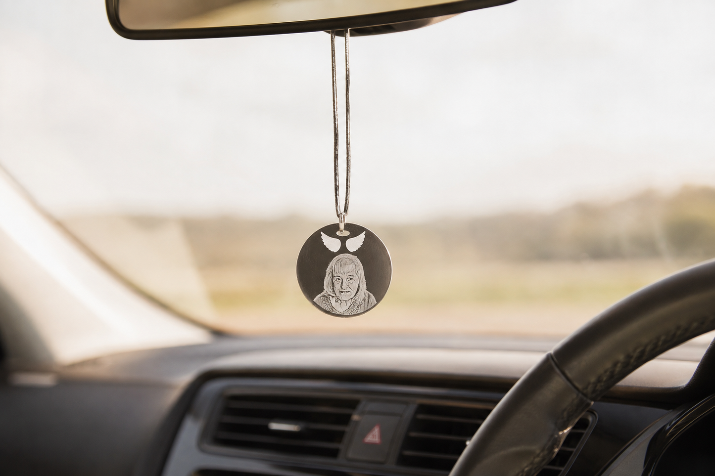 A round black car air freshener pendant with a silver engraved portrait hanging from the rearview mirror inside a car.