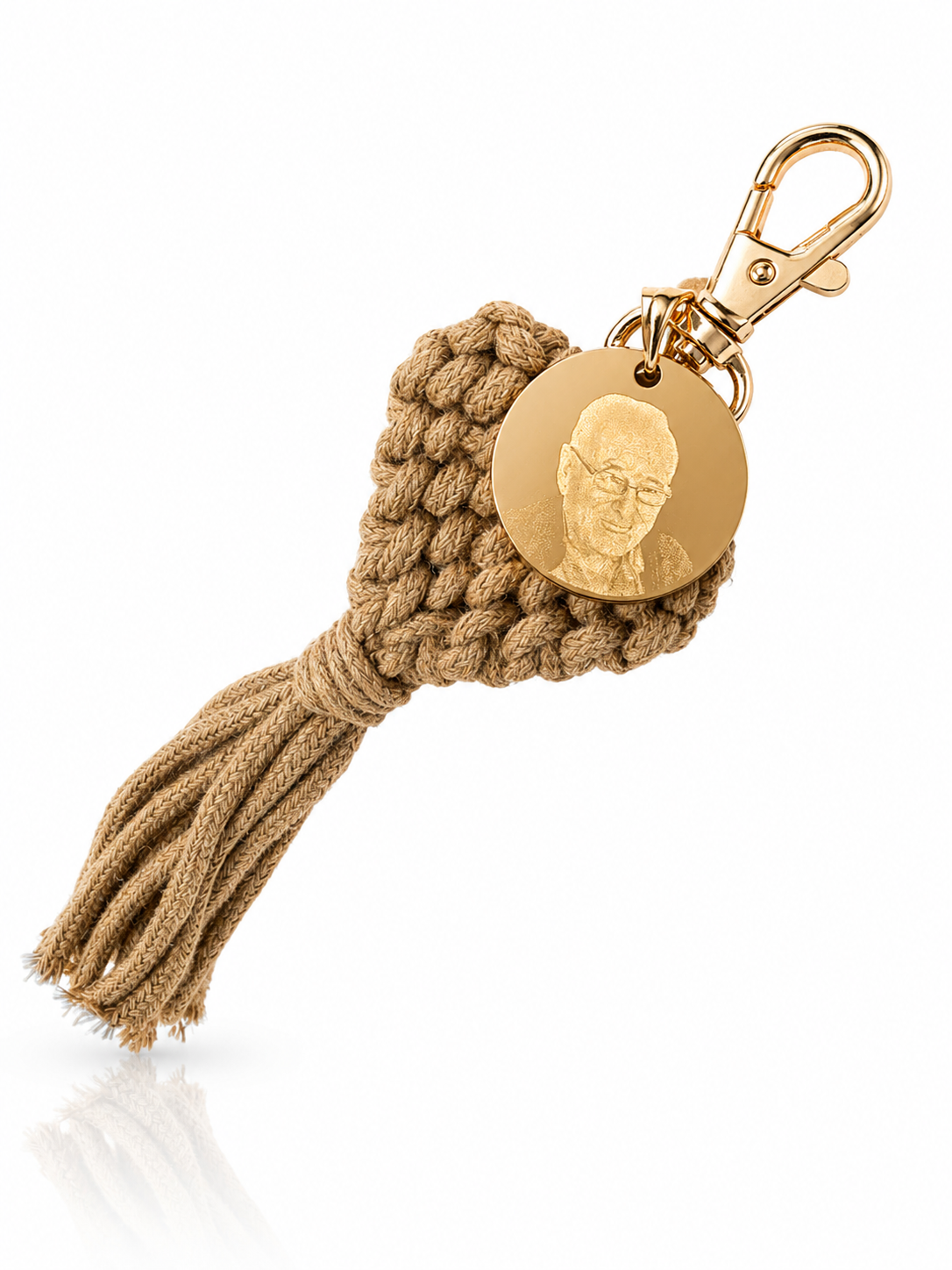 A personalized keychain featuring a gold-toned metal clasp, a round engraved gold disc with a portrait, and a textured tan macrame heart with a matching tassel, shown on a white background with a soft reflection.