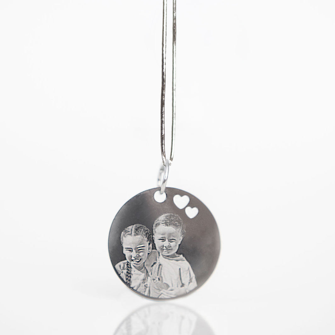 A silver round pendant necklace featuring a custom-engraved black-and-white photo of two children, with two small white heart cutouts near the top, hanging on a thin silver chain against a white background.