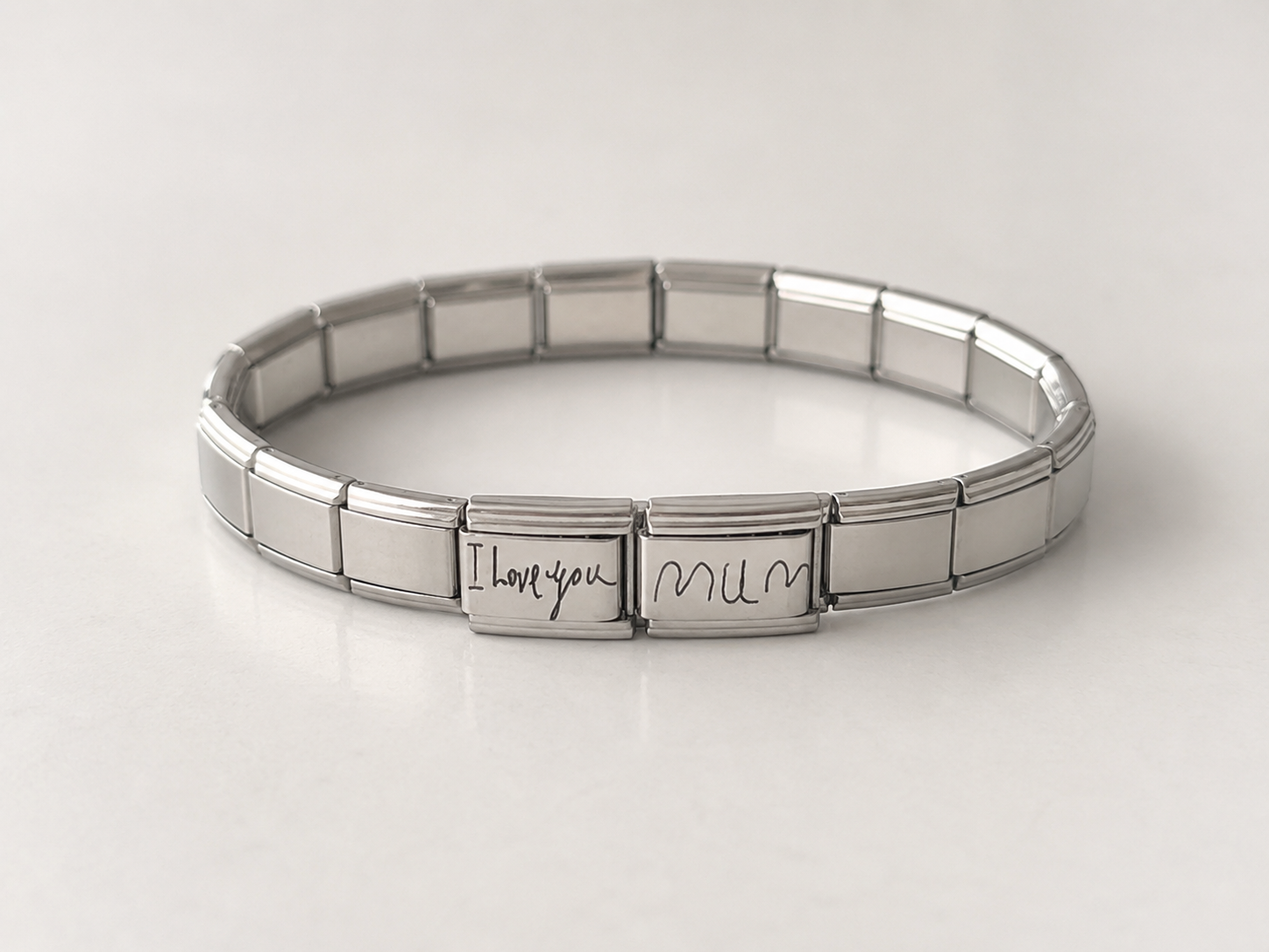 A silver Italian charm link bracelet with a personalized clasp engraved with 'I love you mum' in black script, displayed on a light gray reflective surface.