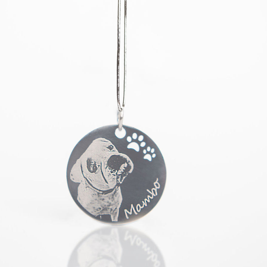 A round black pet memorial pendant with a silver metal wire hanger, featuring a custom engraved dog portrait, paw prints, and the name 'Mambo'.