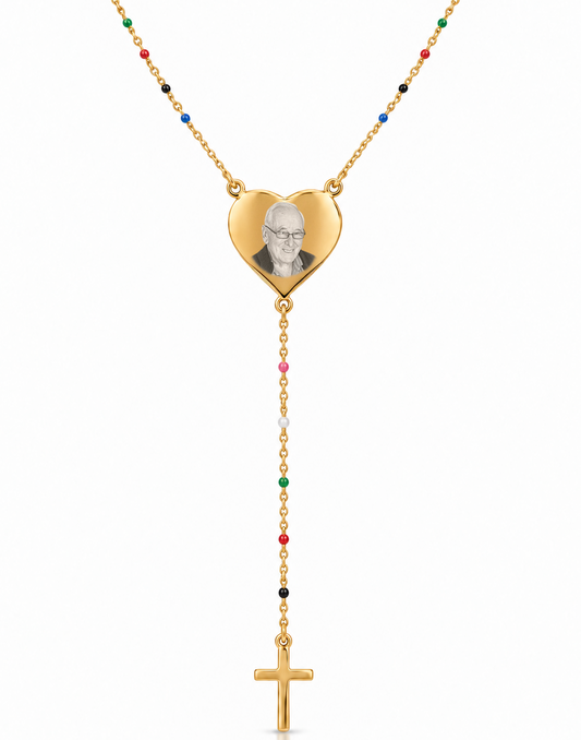 A gold-toned necklace featuring a heart-shaped pendant with a portrait engraving, connected to a rosary-style drop chain with small multicolored beads and a gold cross pendant, shown on a white background.